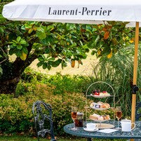 Mobiliario de comedor exterior con un soporte para postres de tres niveles, una mesa con una taza y platillo blancos, una copa de té y una botella de vino bajo un paraguas blanco con la etiqueta 'Laurent-Perrier'.