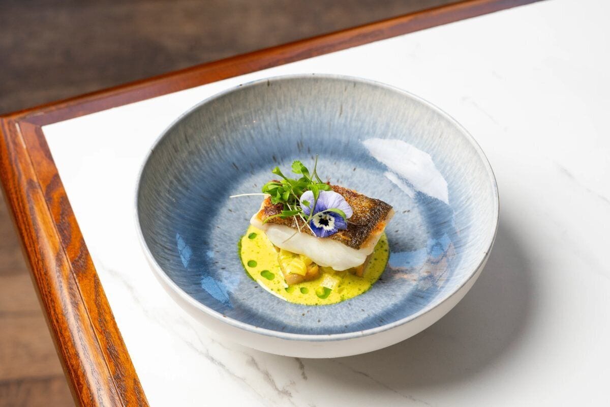 A dish with a piece of fish on a bed of yellow sauce, garnished with greens and a flower, served on a blue ceramic plate.