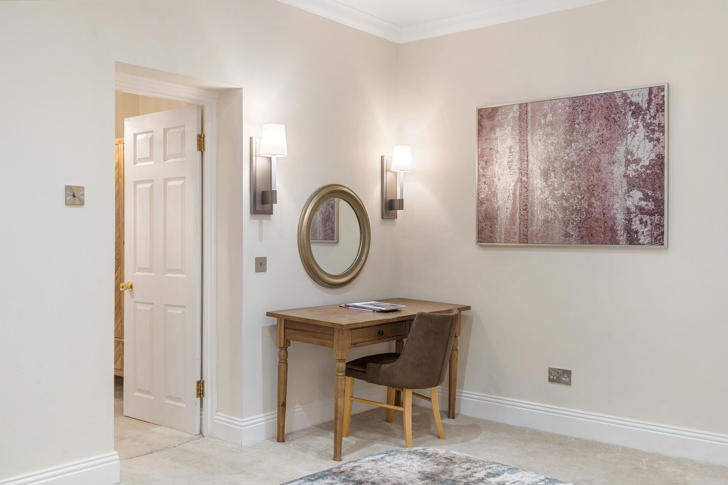 A hallway with a wooden desk, a chair, a round mirror, and two wall-mounted lamps.