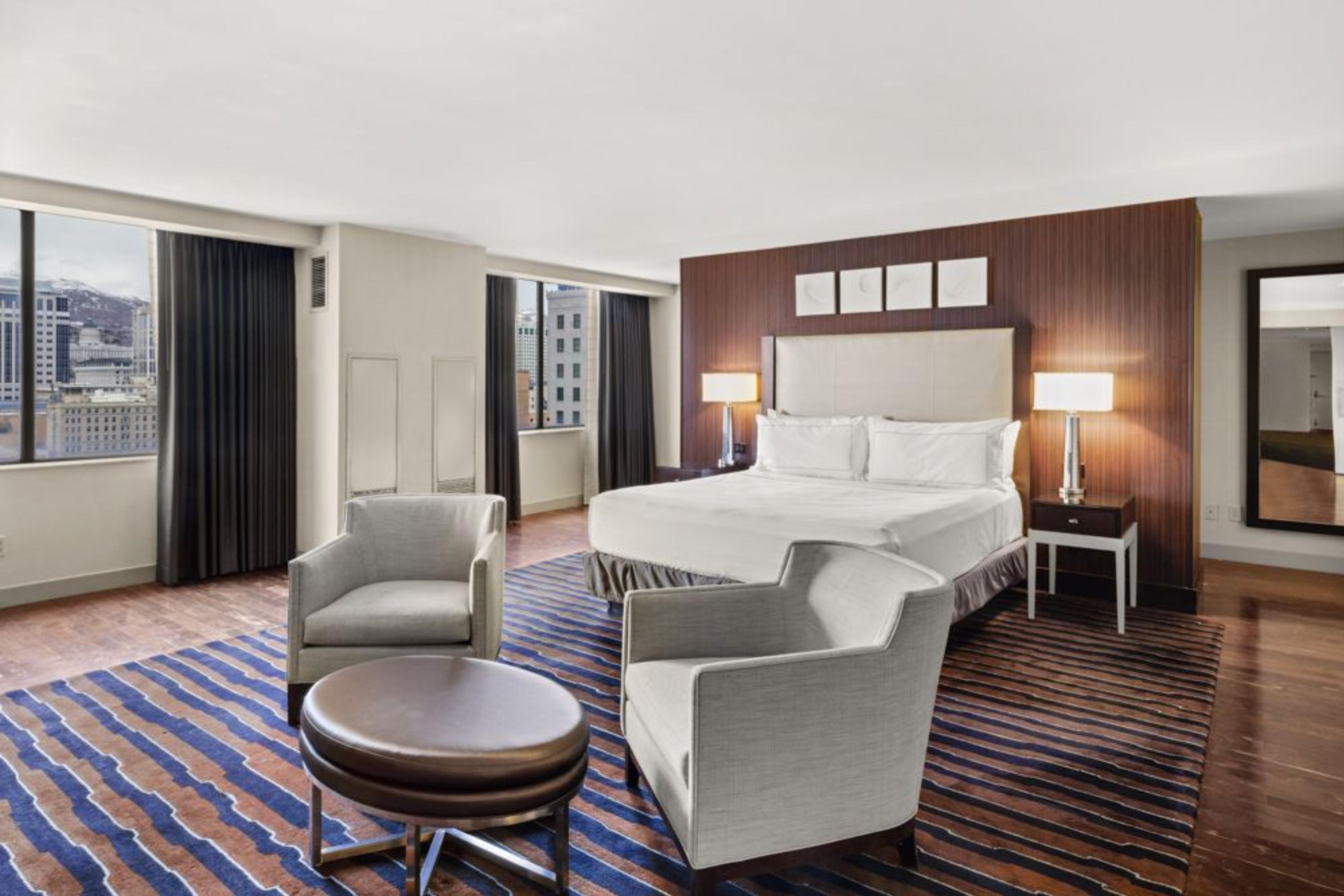 A hotel room with a large bed, two armchairs, a small round table, and a view of the city through the windows.