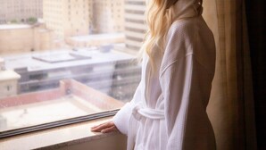A person in a white robe standing by a window with a view of city buildings.