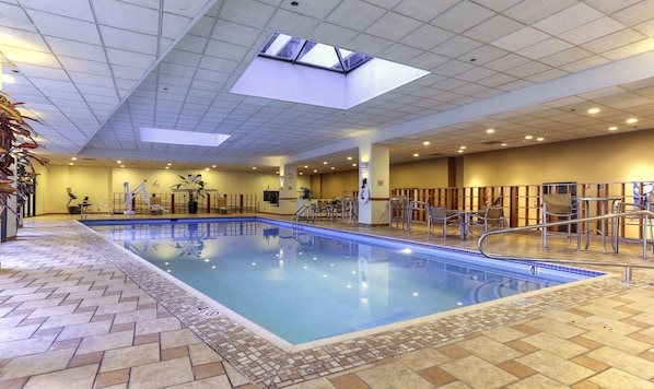 An indoor swimming pool with a tiled floor, a skylight, and a seating area with chairs and tables.