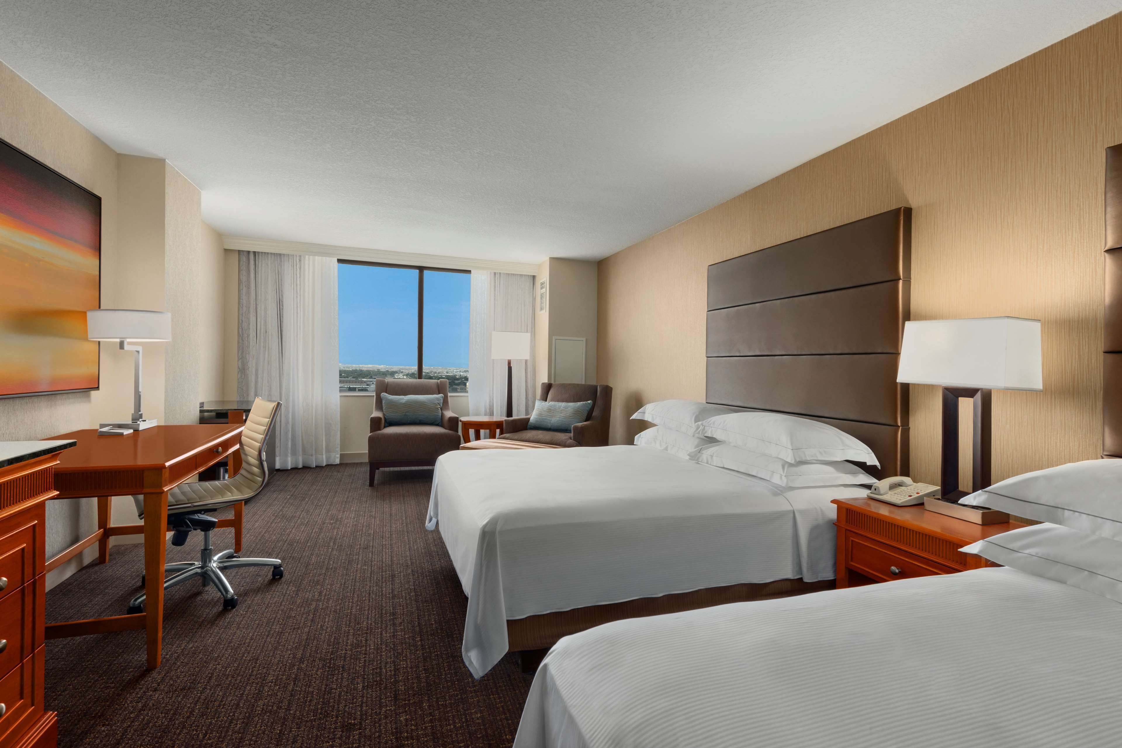 A hotel room with two beds, a desk, a chair, a lamp, and a view of the city.