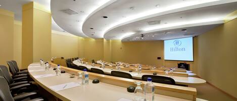 Uma sala de conferências com uma mesa comprida, várias cadeiras, um pódio e uma tela de projeção exibindo o logotipo da Hilton.