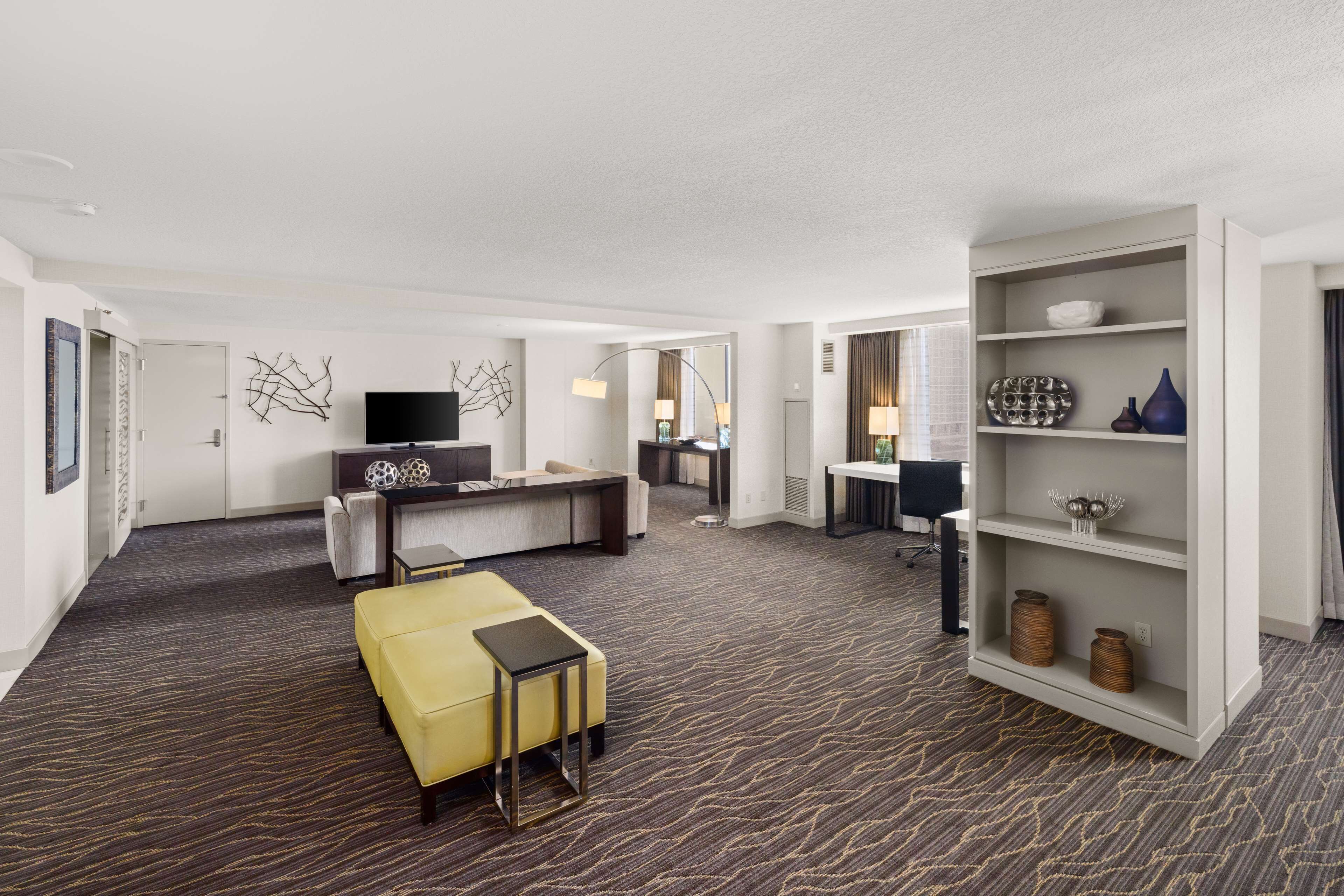 A modern hotel room with a sofa, a yellow ottoman, a desk, and a bookshelf.