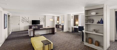 A modern hotel room with a sofa, a yellow ottoman, a desk, and a bookshelf.