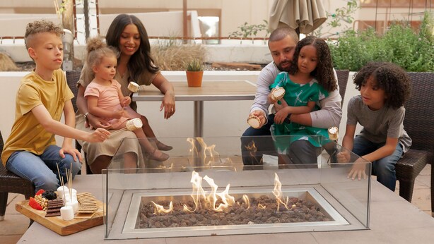 A family around a glass-top table with a fire feature, seated on outdoor furniture.