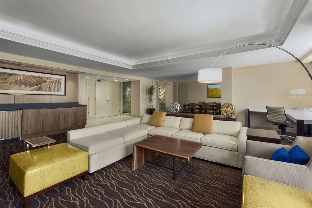 Une chambre d’hôtel moderne, dotée d’un canapé d’angle, d’une petite table basse, d’un tapis à motifs et d’un grand tableau accroché au mur.
