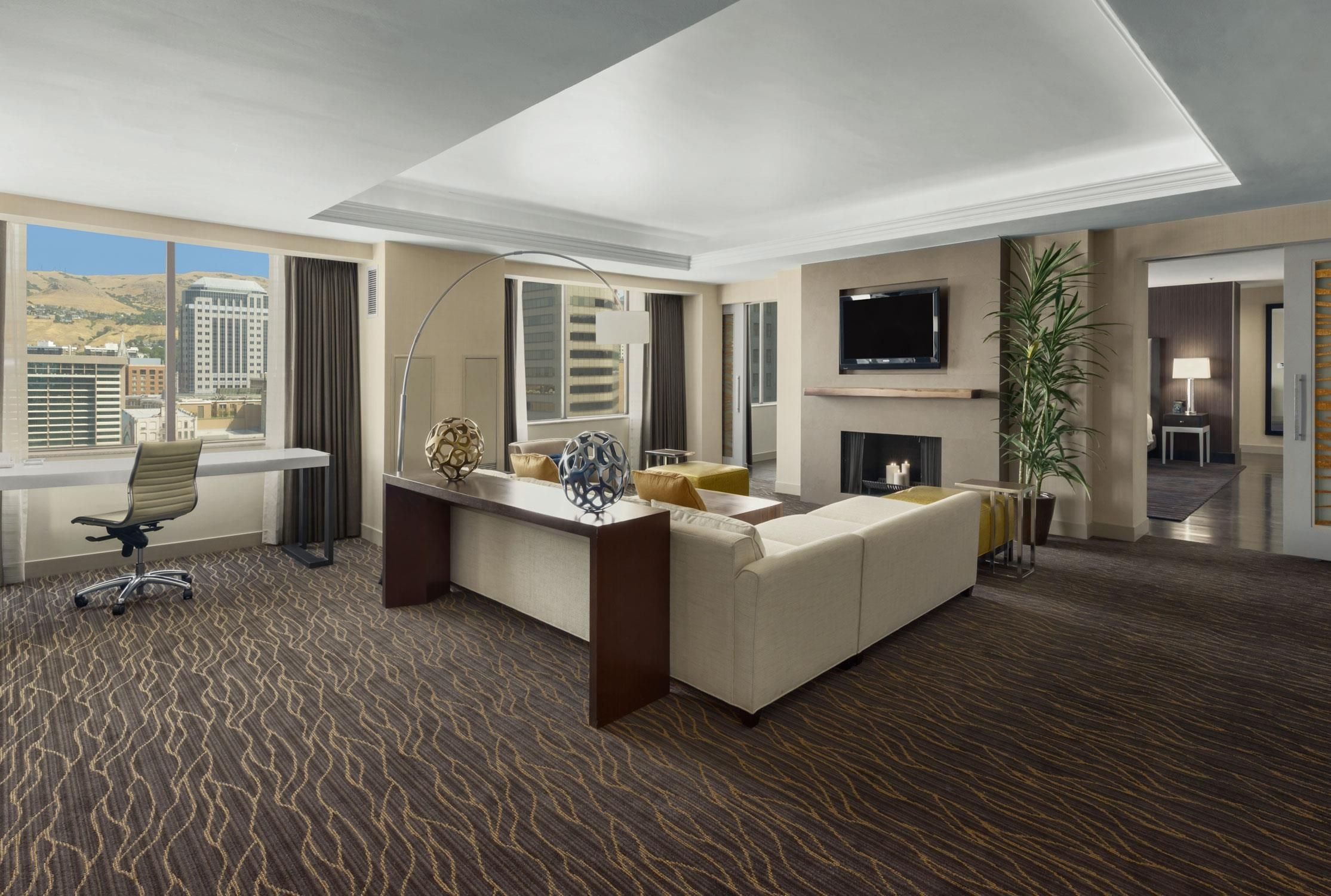 A hotel room with a large bed, a desk, a chair, a TV, a fireplace, and a view of the city.