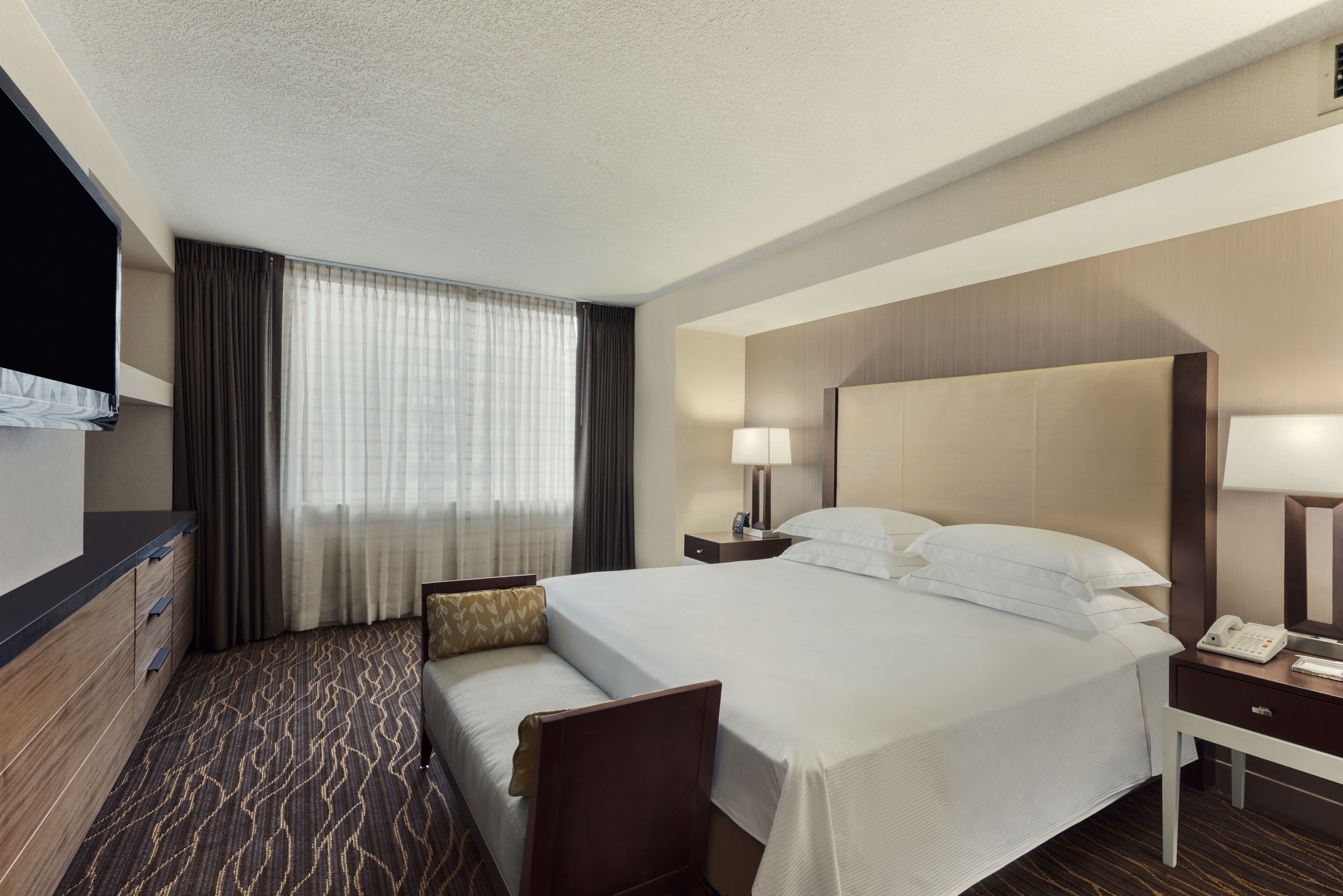 Ein Hotelzimmer mit einem großen Bett, einem Schreibtisch mit Lampe, einem Sessel, einem Fernseher und einem Fenster mit Vorhängen.
