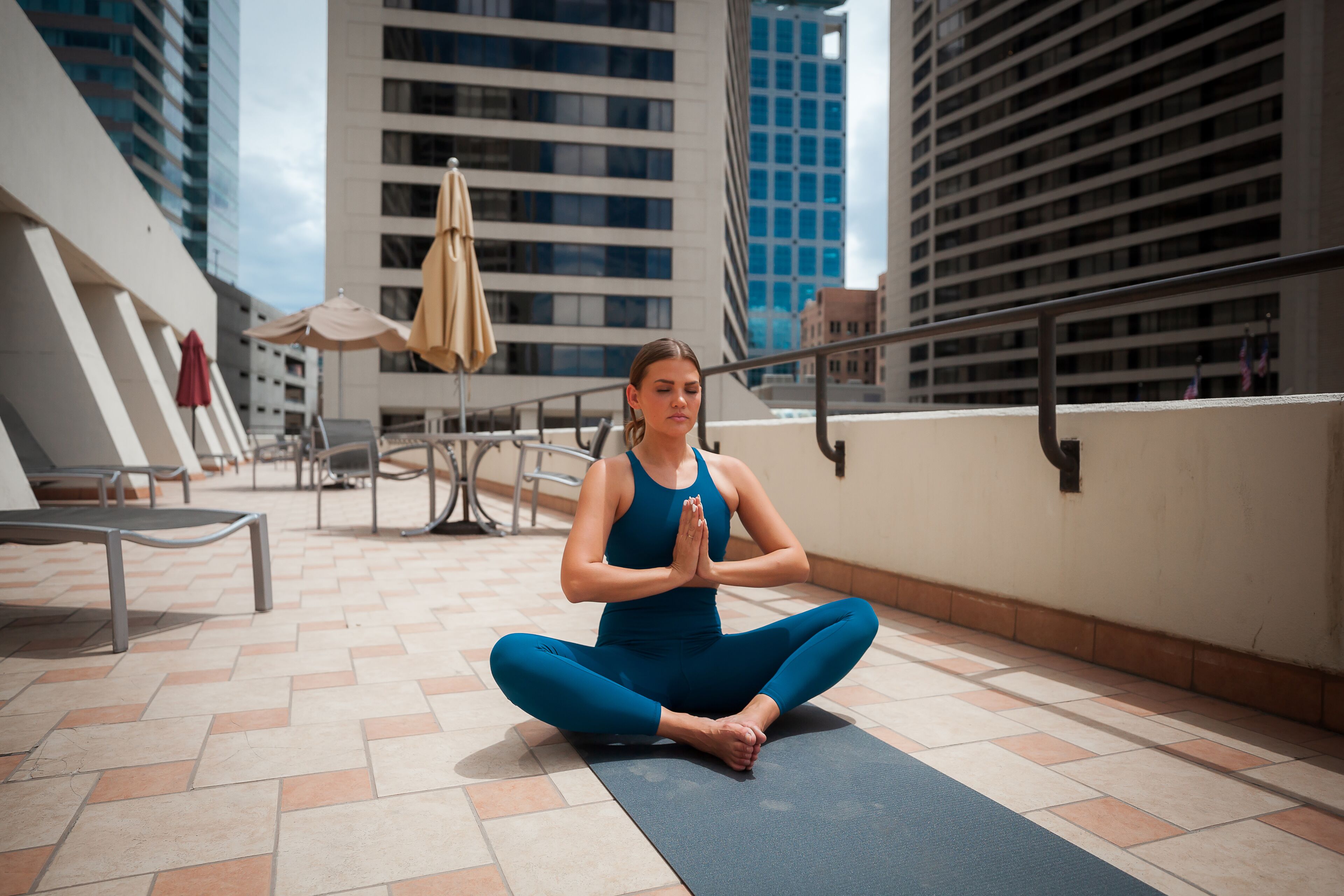 En person sidder på en yogamåtte i et udendørs område med gelænder og bygninger i baggrunden.