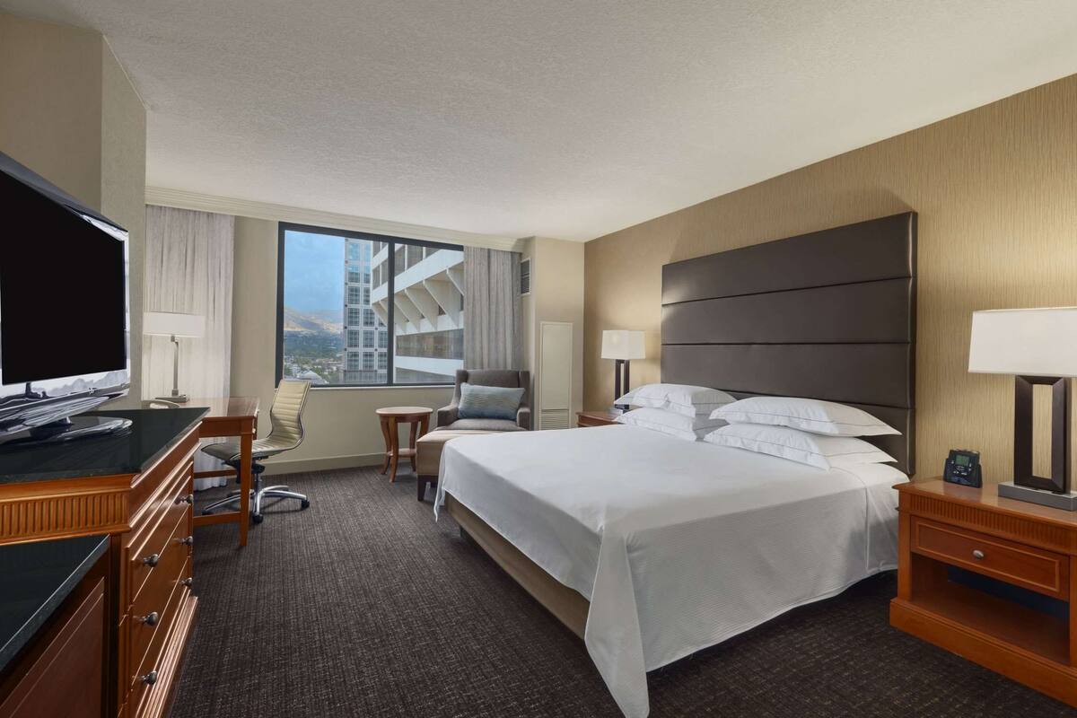 Une chambre d’hôtel avec un grand lit, un bureau avec une télévision, une chaise et une vue sur un immeuble par la fenêtre.