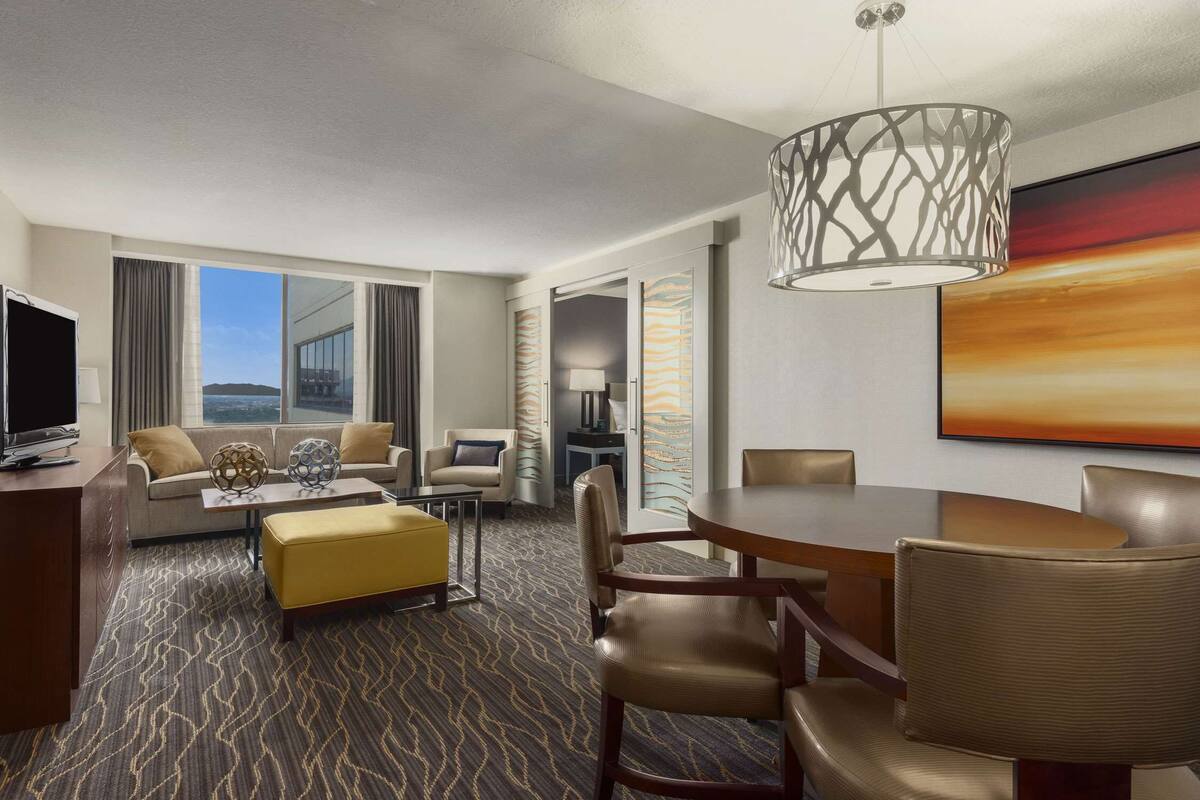 Une chambre d’hôtel moderne équipée d’un canapé, d’un pouf, d’une table à manger, de chaises, d’un téléviseur et d’une grande fenêtre donnant sur la mer.