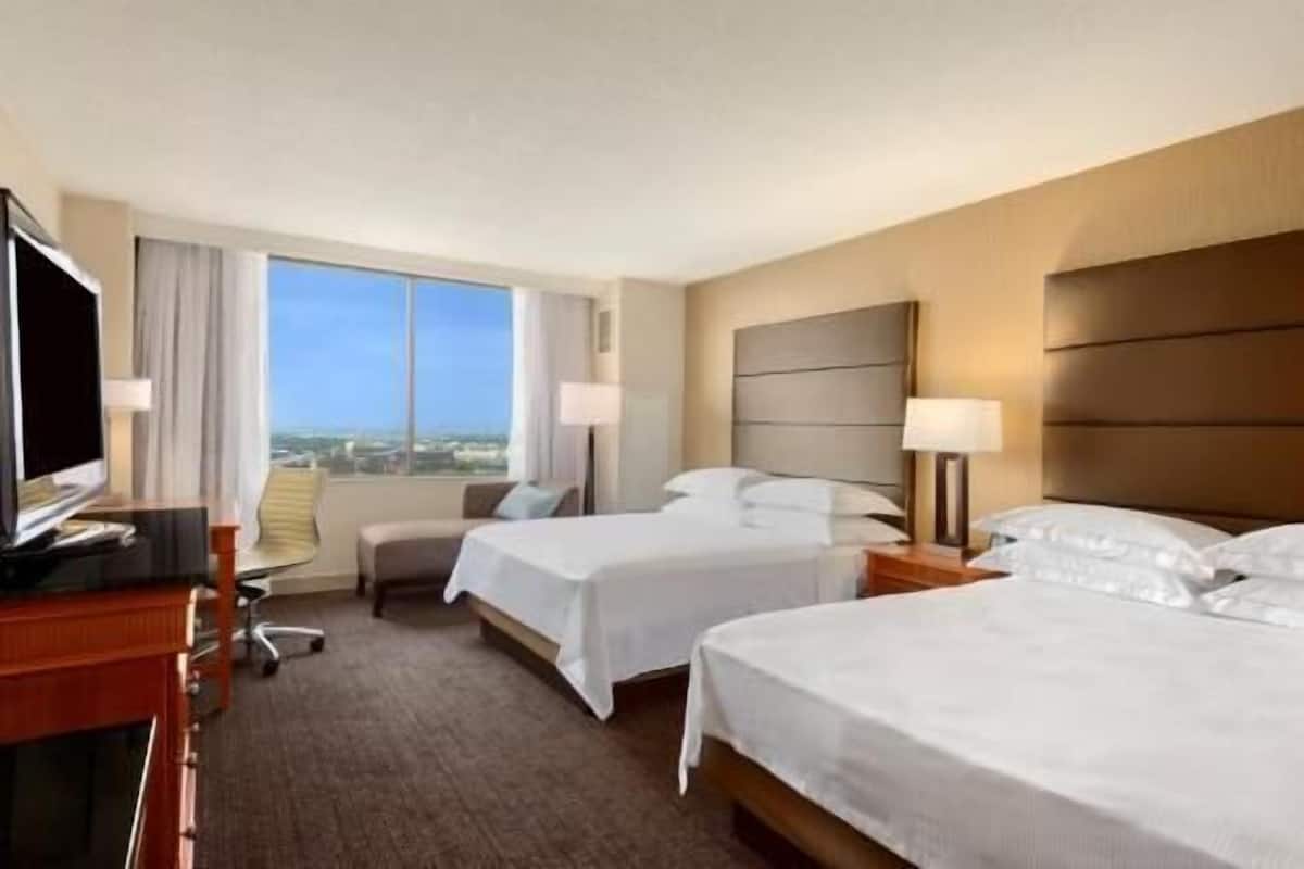 Habitación de hotel con dos camas, un escritorio con computadora, una silla y vistas a la ciudad.