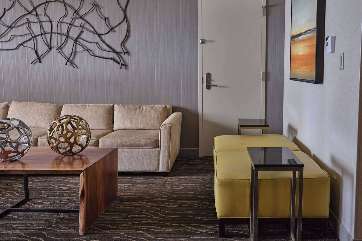 Un salon moderne comprenant un canapé, une table basse et un pouf, un élément décoratif mural et une porte surmontée d’un tableau.