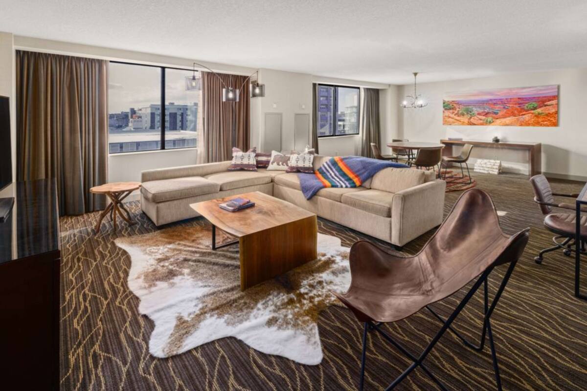 Une chambre d’hôtel moderne, dotée d’un canapé d’angle, d’une table basse en bois, d’un fauteuil en cuir et d’une grande fenêtre donnant sur la ville.