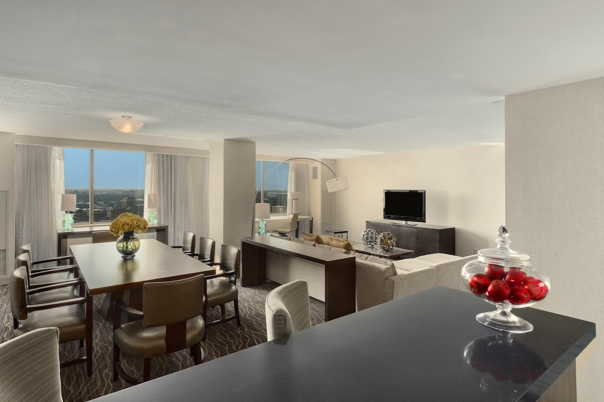 Une chambre d’hôtel comprenant un coin repas, un canapé, une télévision et un saladier en verre rempli de pommes.
