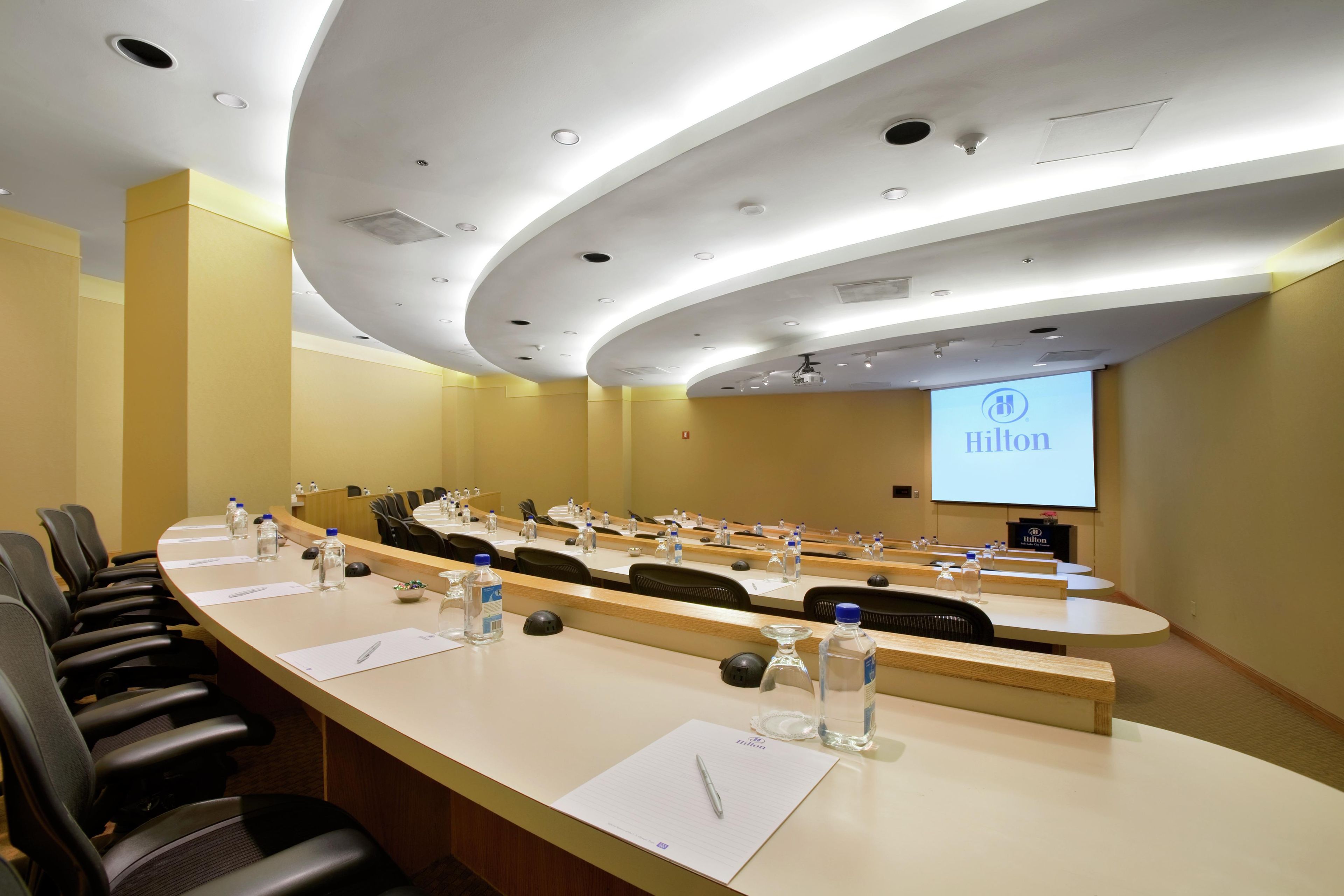 Un salón de conferencias con una larga mesa, sillas, botellas de agua y una pantalla proyectora mostrando el logo de Hilton.