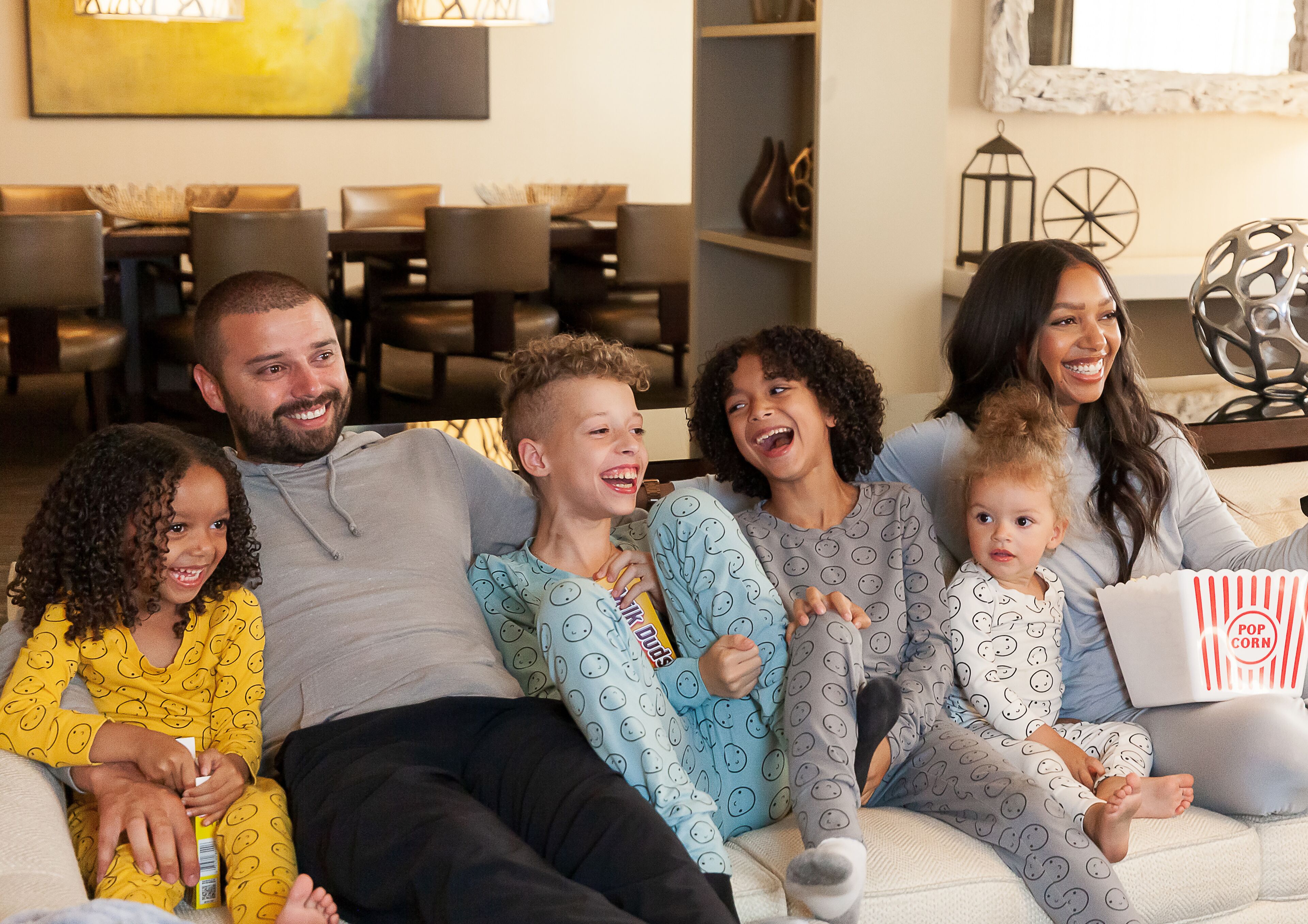 A family sitting on a couch in a living room with a dining area in the background.