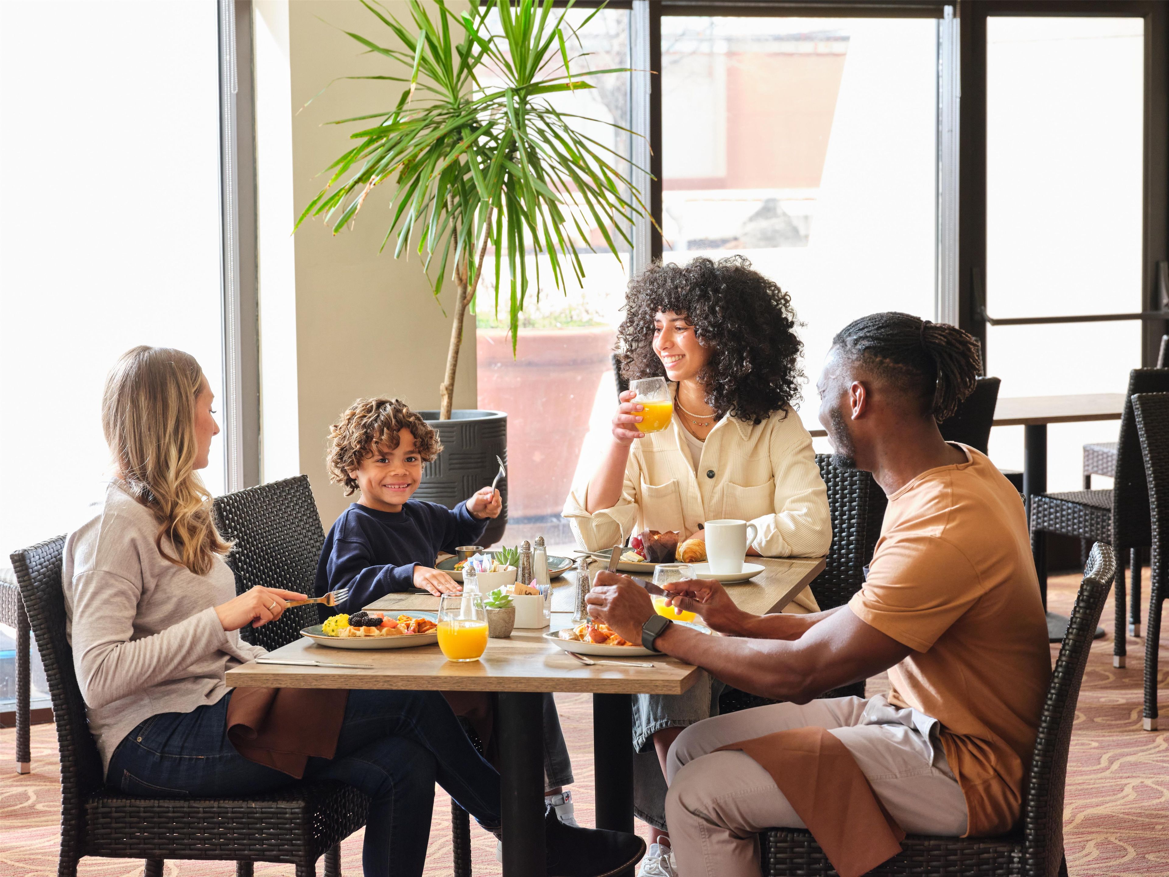 A group of people dining at an outdoor table with a potted plant and a large window in the background.