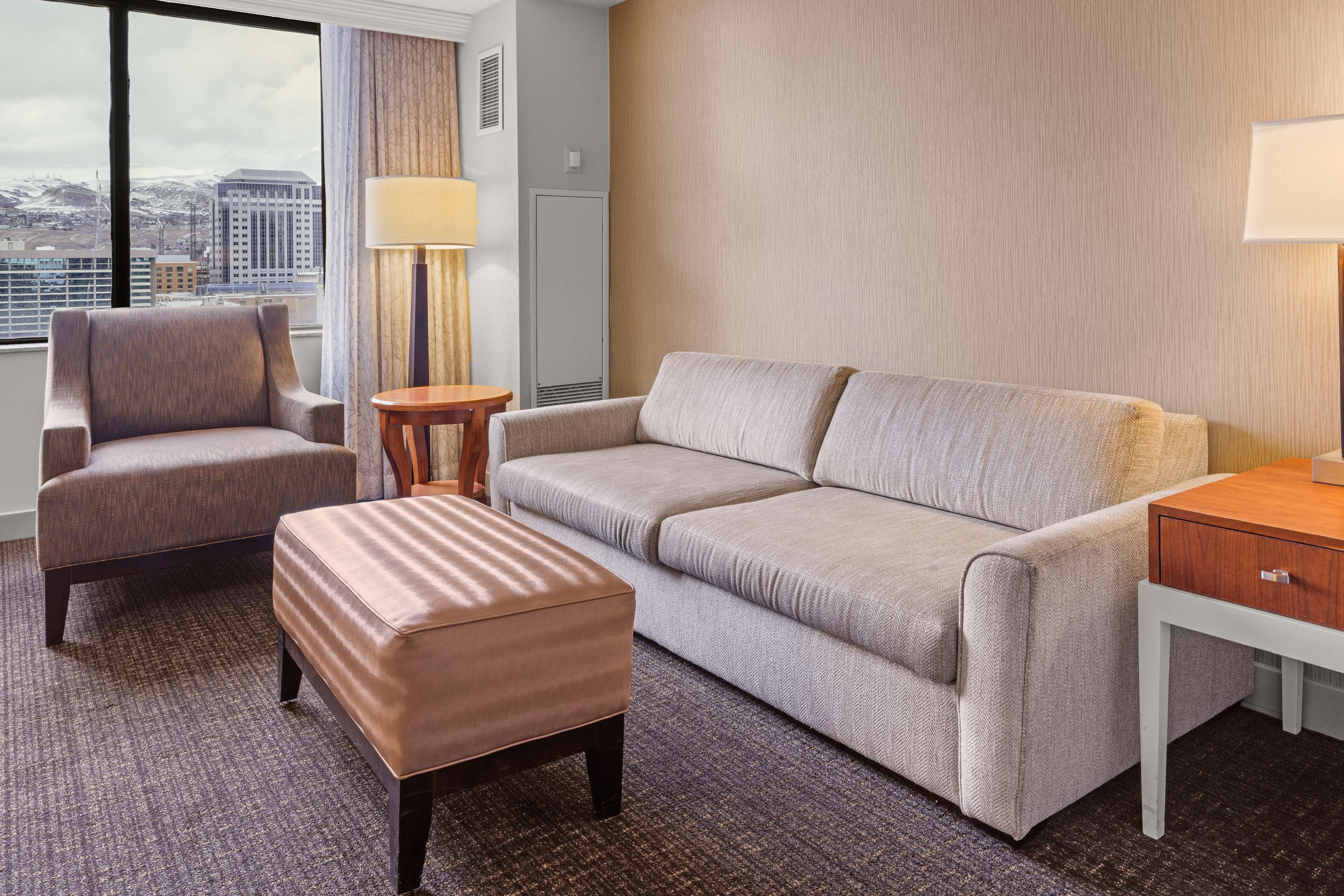 A hotel room with a grey sofa, a matching armchair, a wooden side table, and a small ottoman.