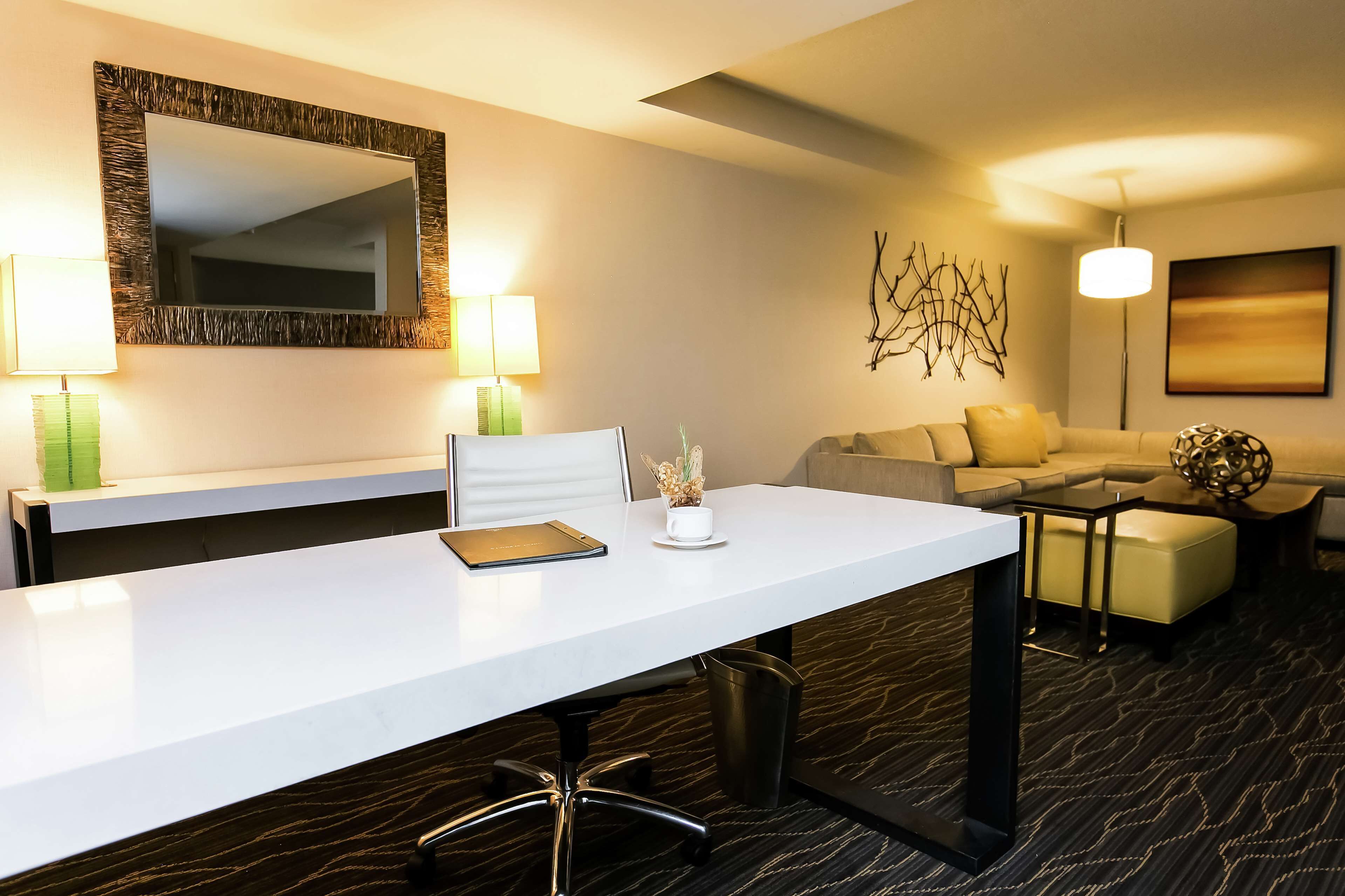 Une chambre d’hôtel avec un bureau blanc, deux lampes, un canapé, une table basse et un élément décoratif mural.