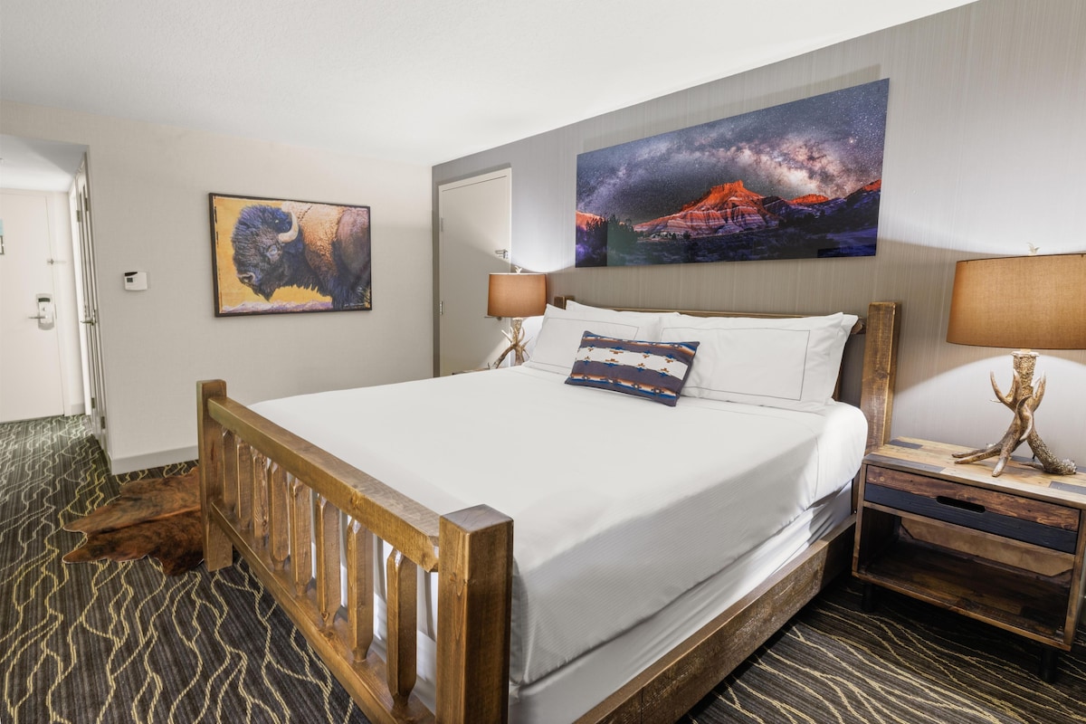 Une chambre d’hôtel avec un grand lit, une table de chevet, une lampe, un tableau représentant un paysage de montagne et une porte.