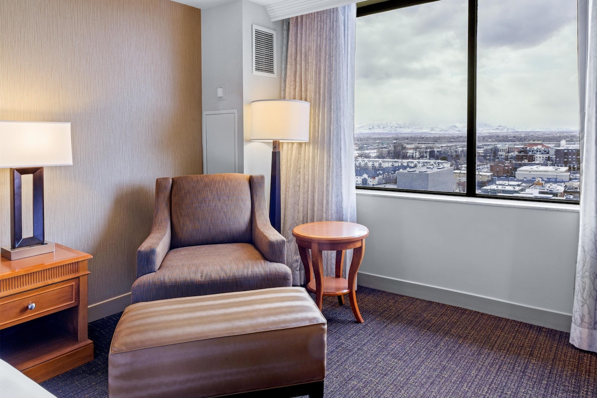 Une chambre d’hôtel avec une grande fenêtre, une chaise, une petite table et une lampe.