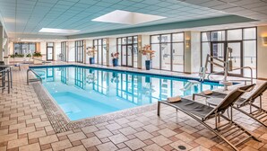 An indoor swimming pool with clear glass walls, lounge chairs, and potted plants.
