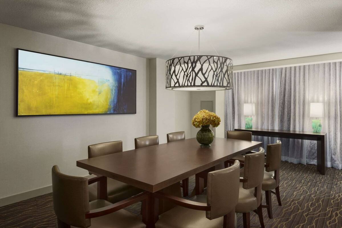 Une salle à manger d’hôtel avec une longue table, de nombreuses chaises, un vase rempli de fleurs jaunes, un lustre moderne et un grand tableau abstrait accroché au mur.