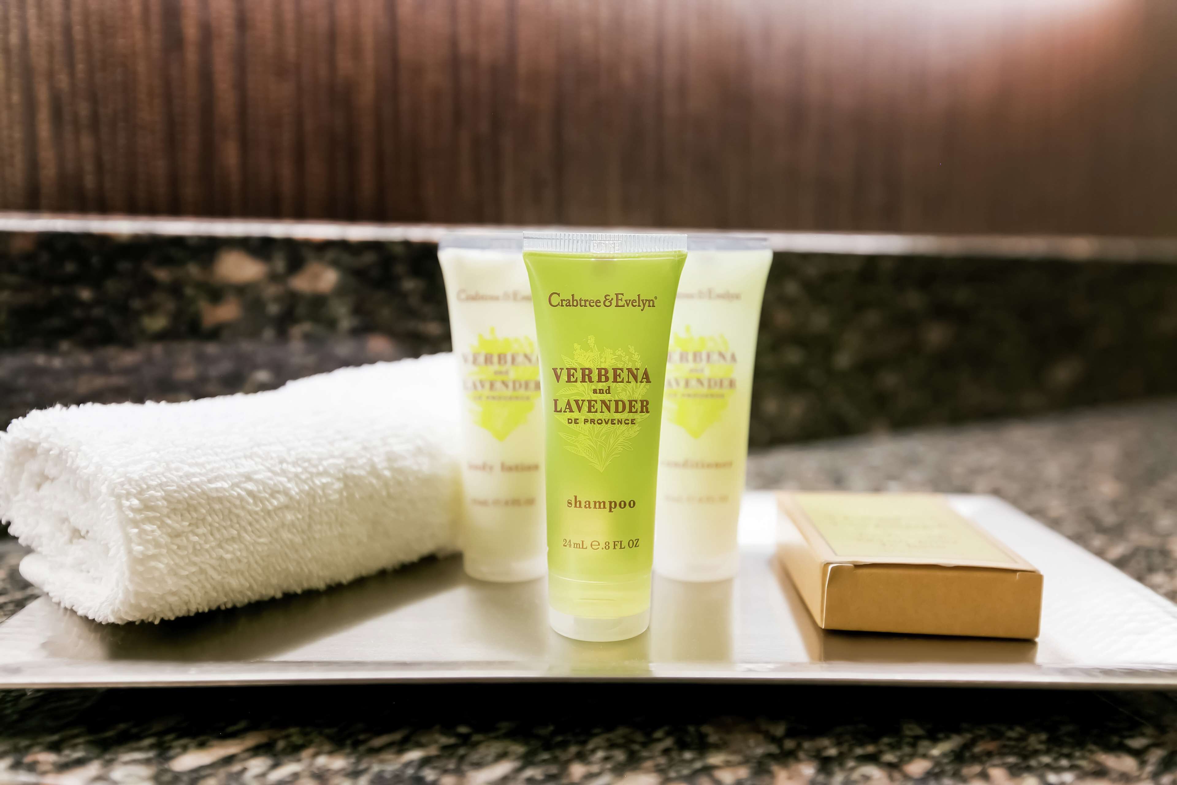 A bathroom counter with a white towel, three bottles of Crabtree & Evelyn Verbenia Lavender shampoo, and a bar of soap.