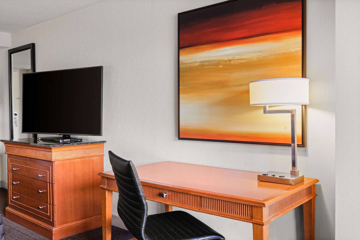 Une chambre d’hôtel avec un bureau en bois, un téléviseur posé sur une commode, une lampe et un grand tableau abstrait accroché au mur.