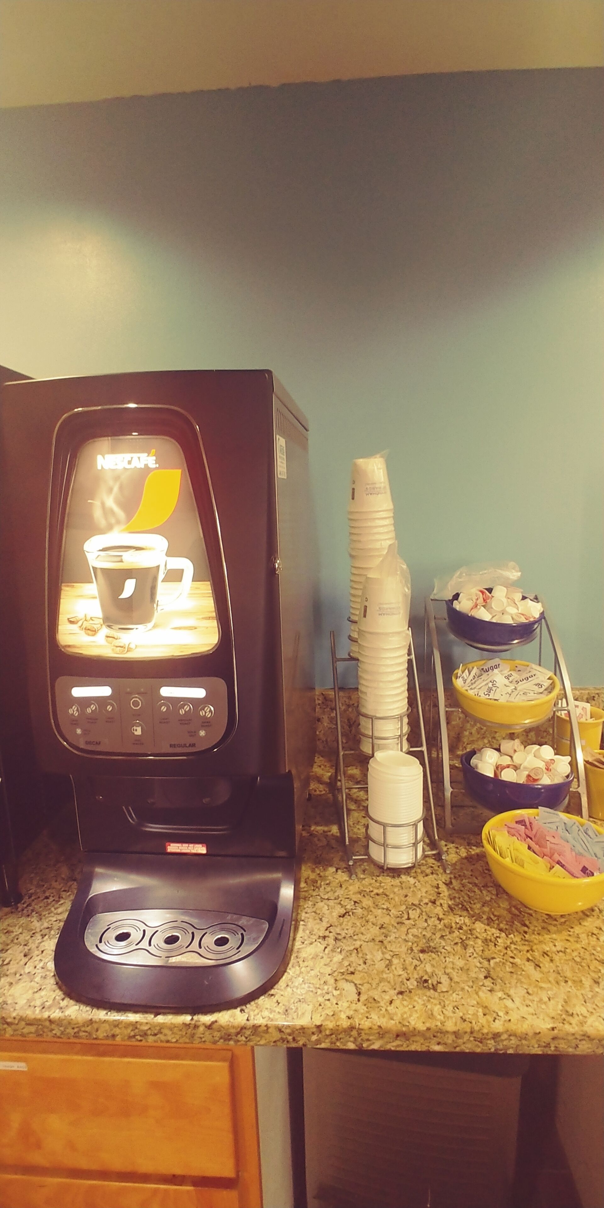 A coffee machine with a digital display, stacked plastic cups, and bowls of sugar and condiments on a kitchen counter.