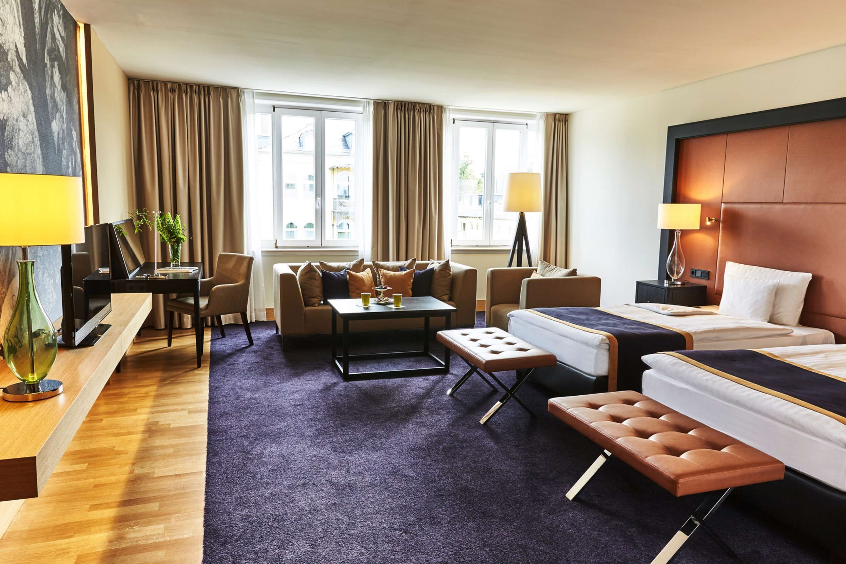 A hotel room with a bed, a sofa, a desk, and a chair. There are two windows with curtains, a lamp, and a plant.