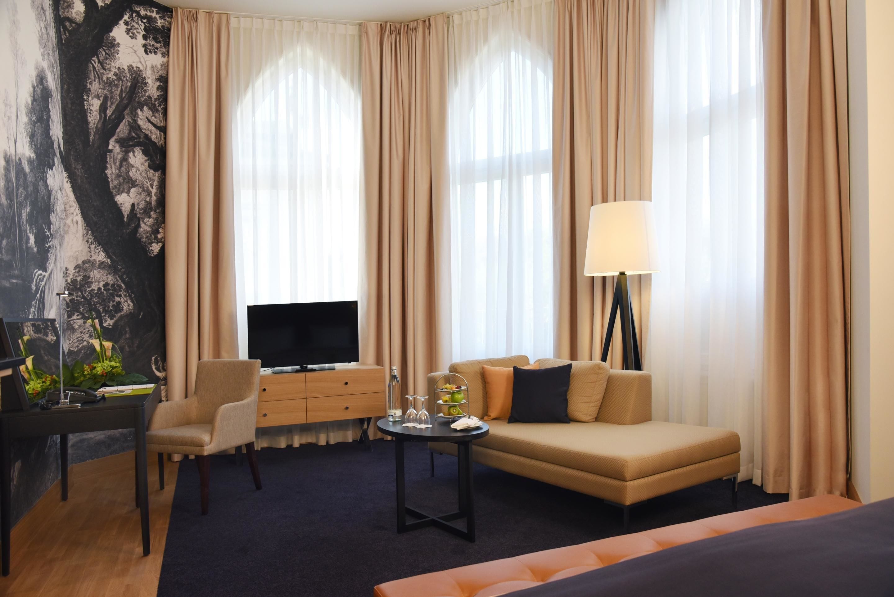 A hotel room with a sofa, armchair, TV, and a desk with a lamp.
