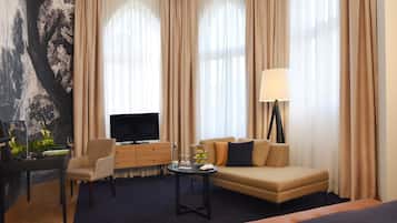A hotel room with a sofa, armchair, TV, and a desk with a lamp.