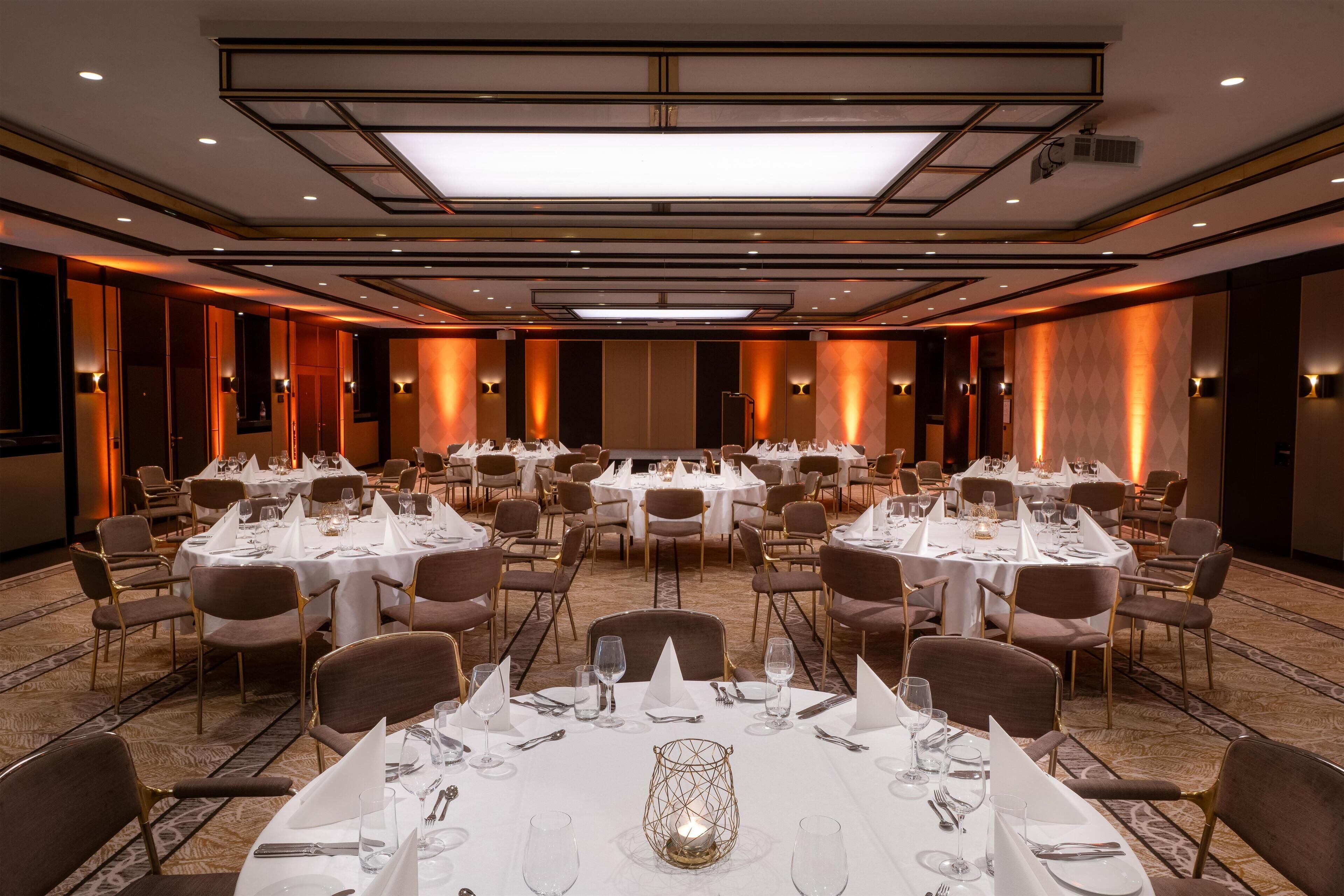 Un salón de banquetes con varias mesas redondas dispuestas para comer, equipado con manteles blancos, sillas y cubertería.
