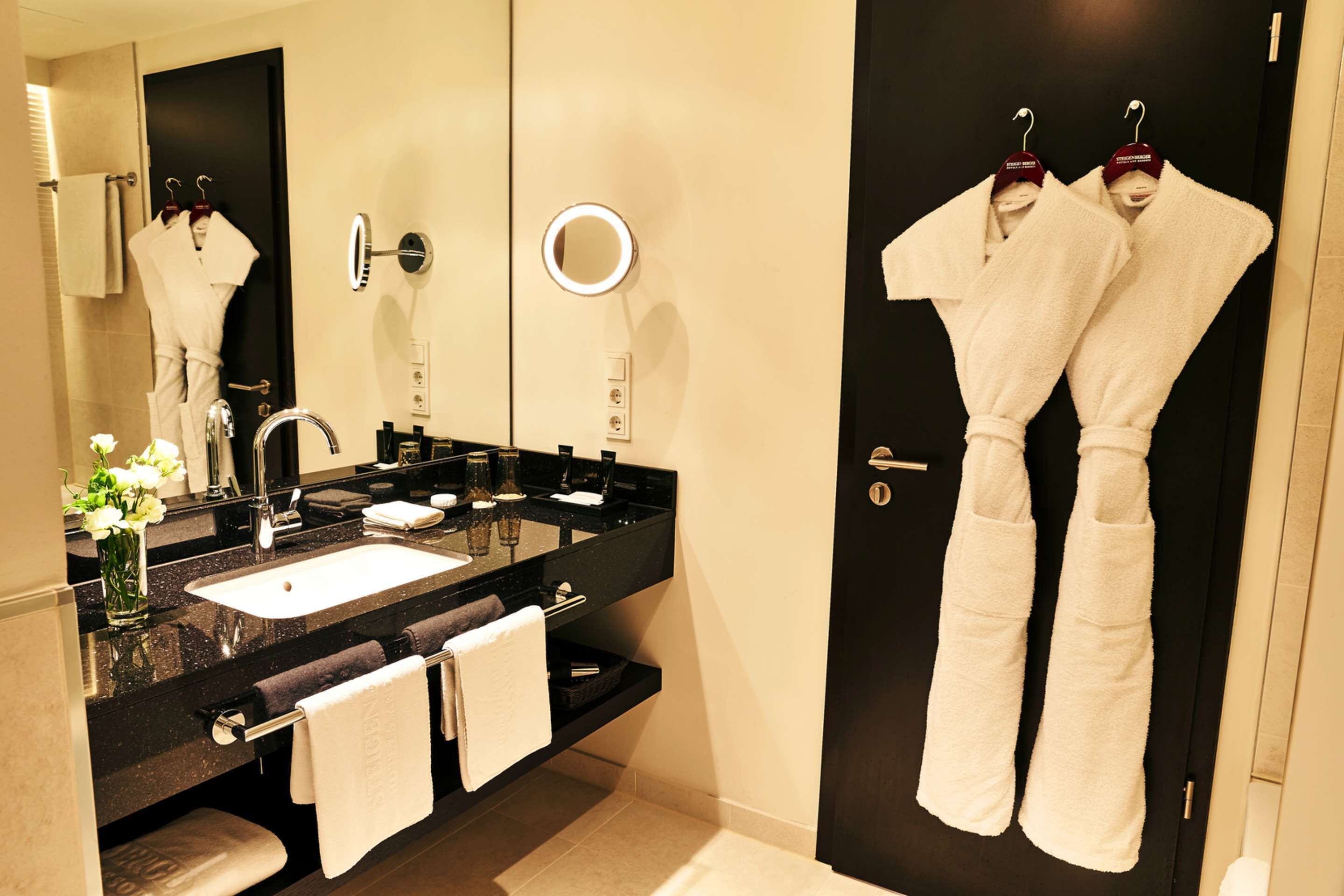 A modern bathroom with a black countertop, two sinks, a large mirror, and a robe rack with towels.