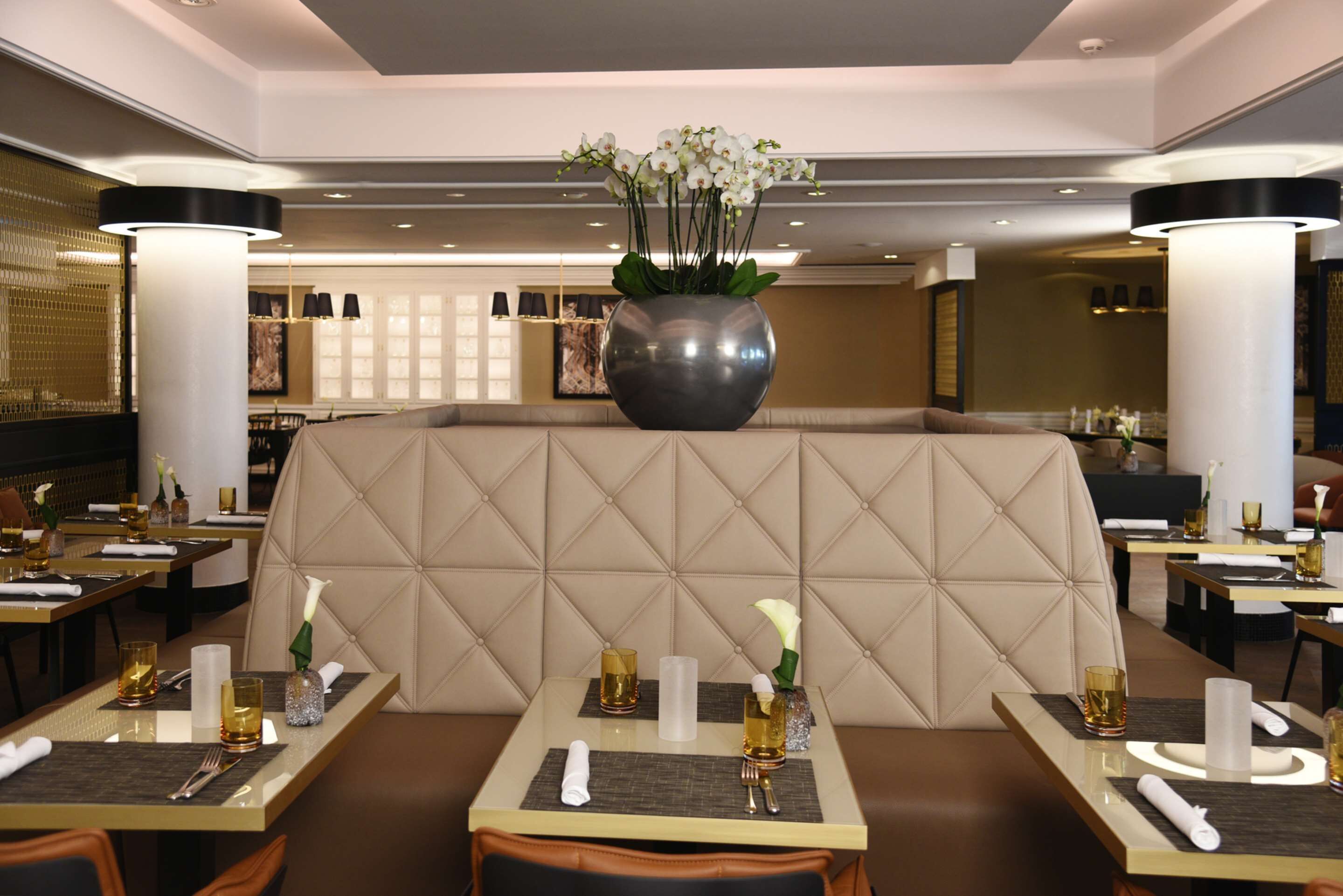 Ein Restaurant mit einer zentralen Sitzgruppe, die von einer großen Vase mit weißen Blumen umgeben ist, und Tischen mit ordentlich gedecktem Geschirr.