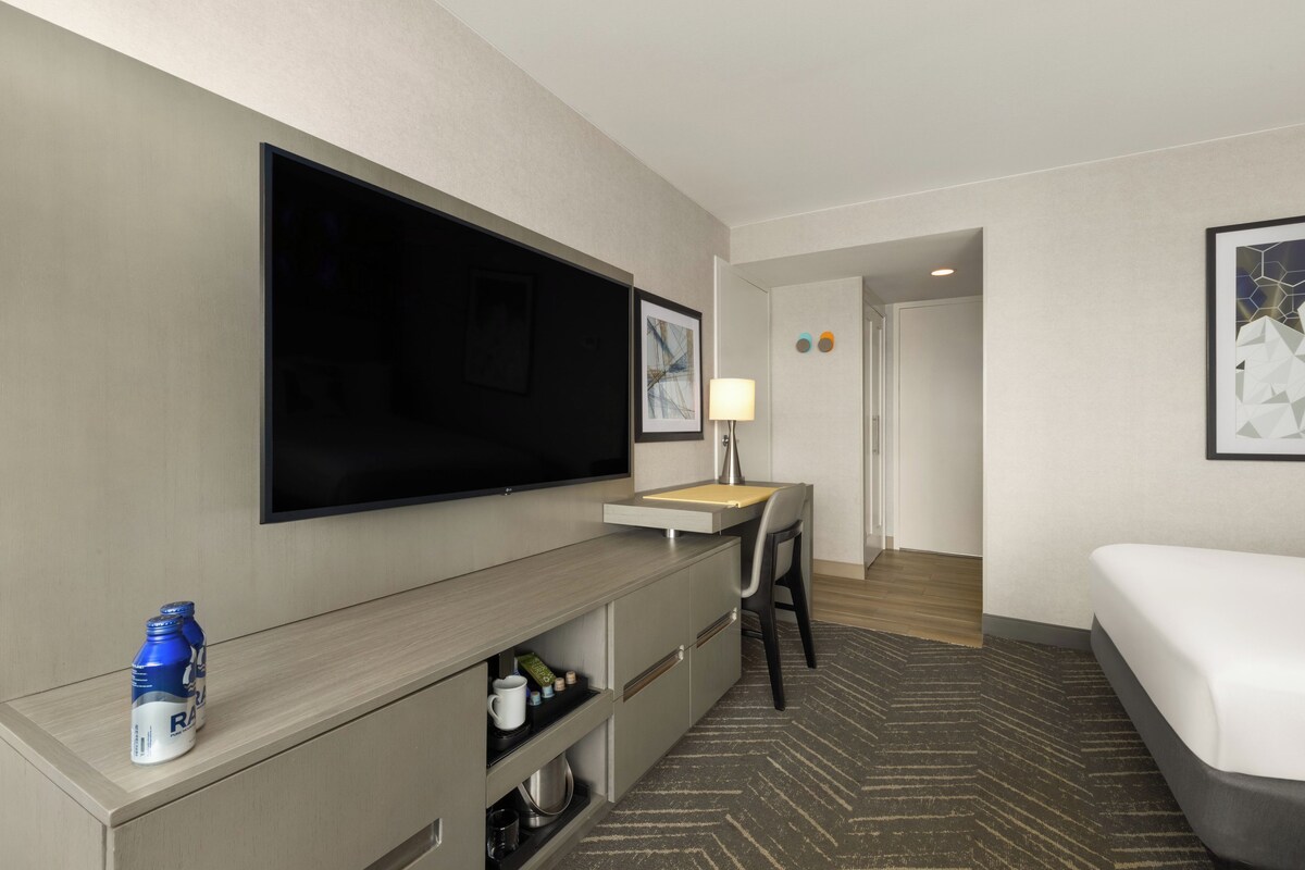 A hotel room with a flat-screen TV, a desk with a lamp, a bed, and a minibar.