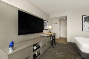 A hotel room with a flat-screen TV, a desk with a lamp, a bed, and a minibar.
