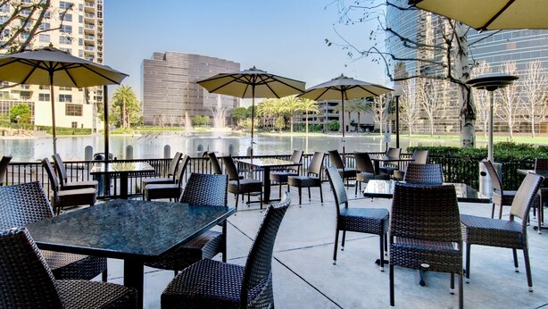 An outdoor patio area with tables and chairs, umbrellas, and a view of a water body and buildings. - voco Orange County - Airport by IHG (Santa Ana)