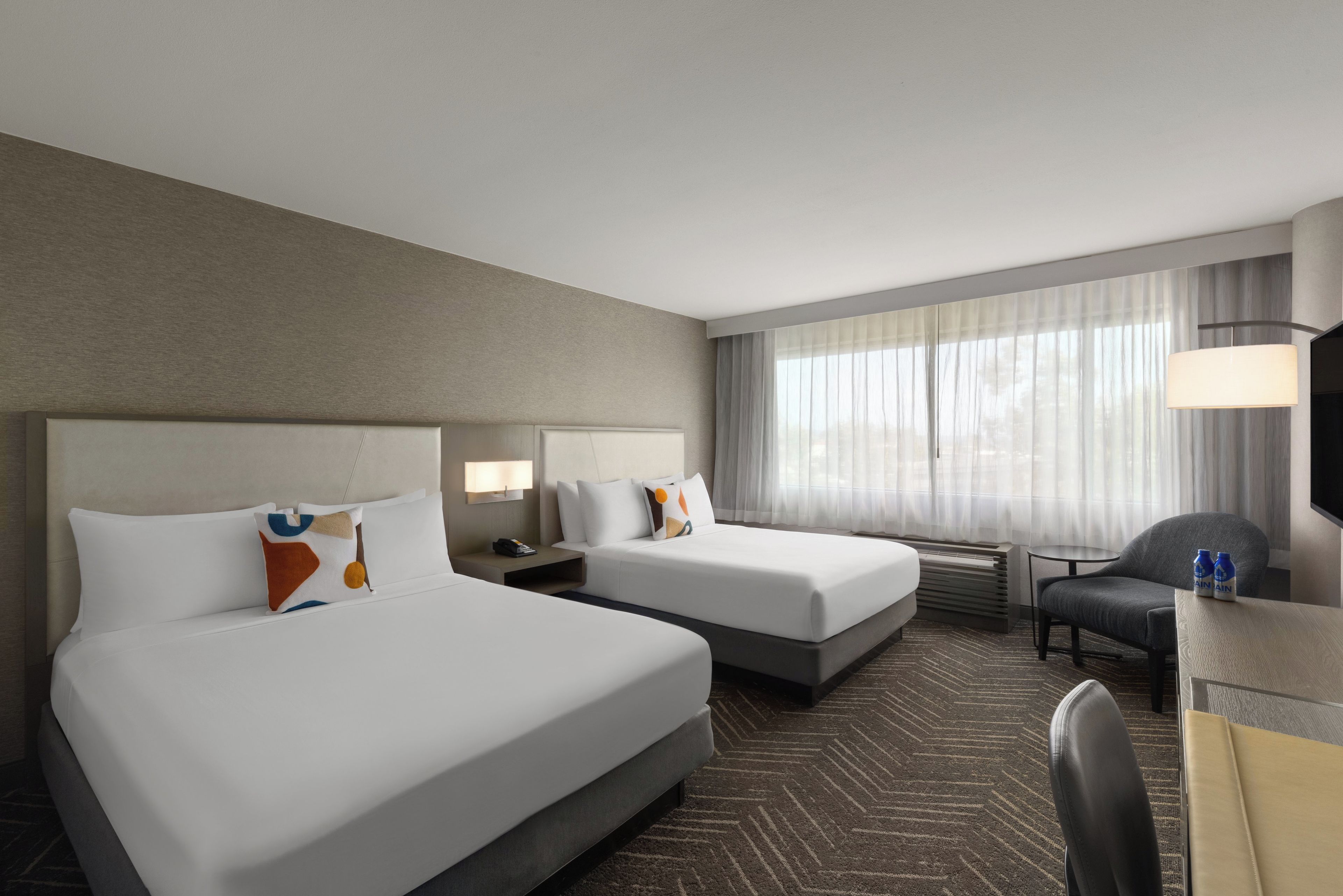 A hotel room with two beds, a desk, a chair, and a large window with curtains.