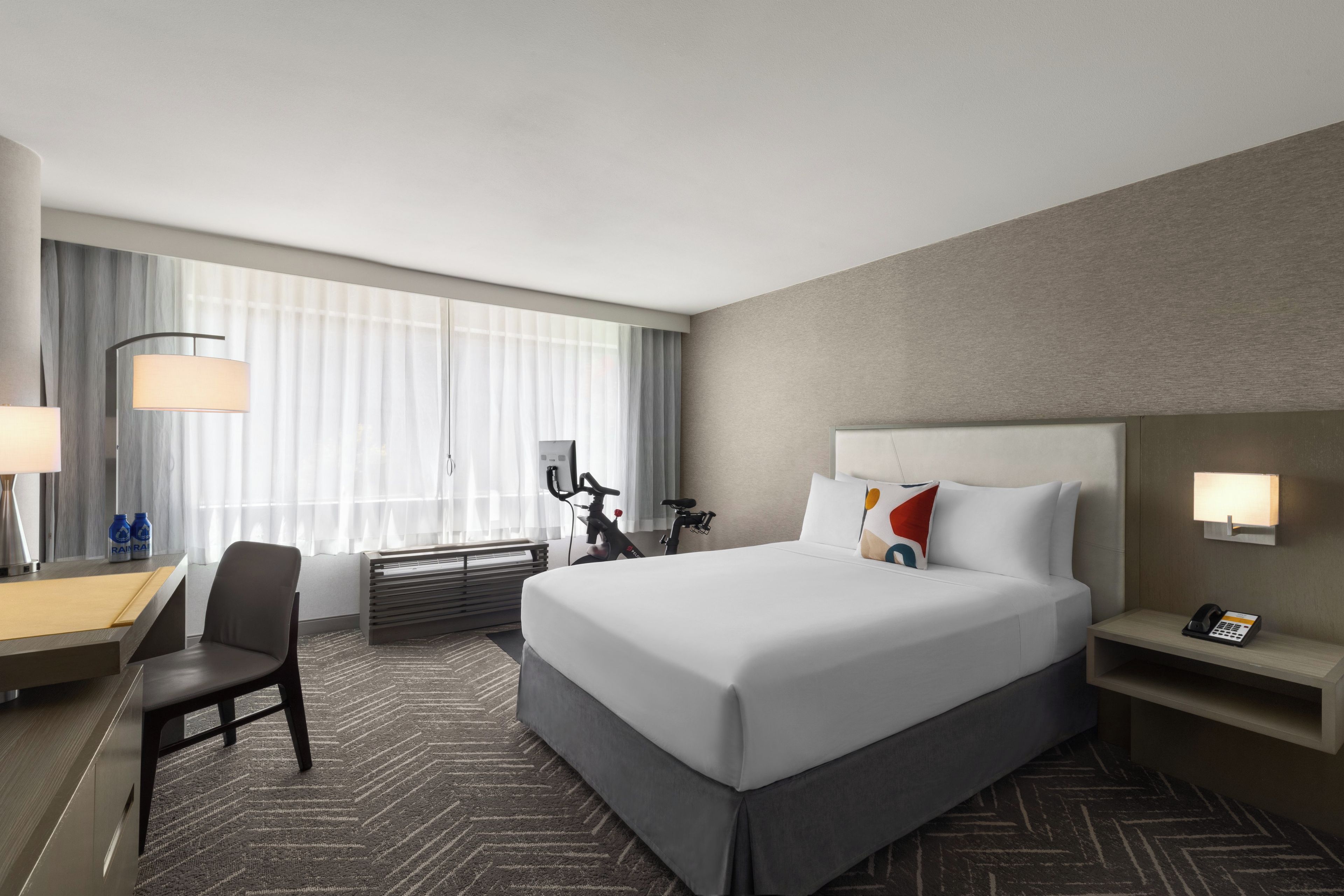 A hotel room with a large bed, a desk, a chair, a bicycle, and a phone on the nightstand.