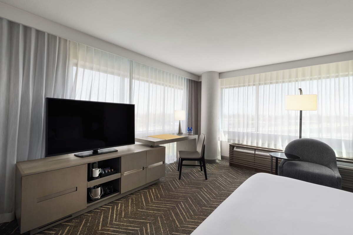 A hotel room with a flat-screen TV, a desk with a lamp, a chair, and a bed.