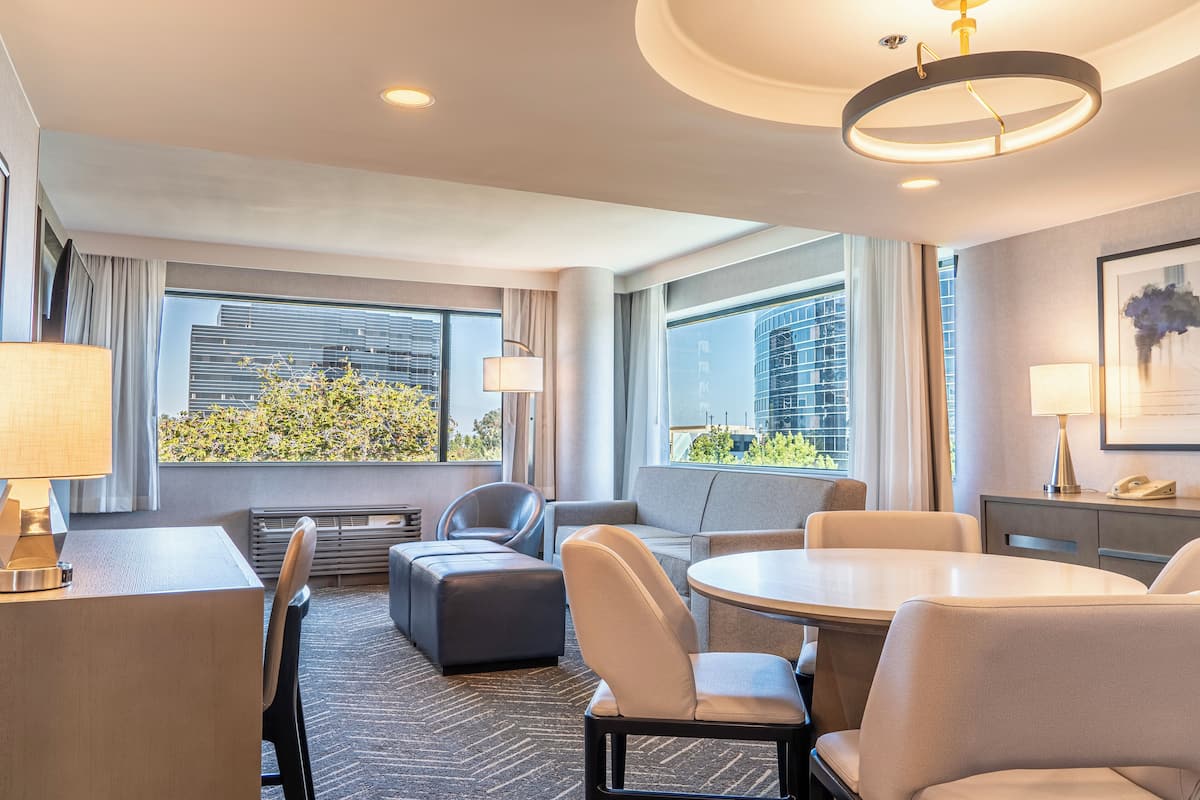 A modern hotel room with a dining area, seating, and large windows offering a city view.