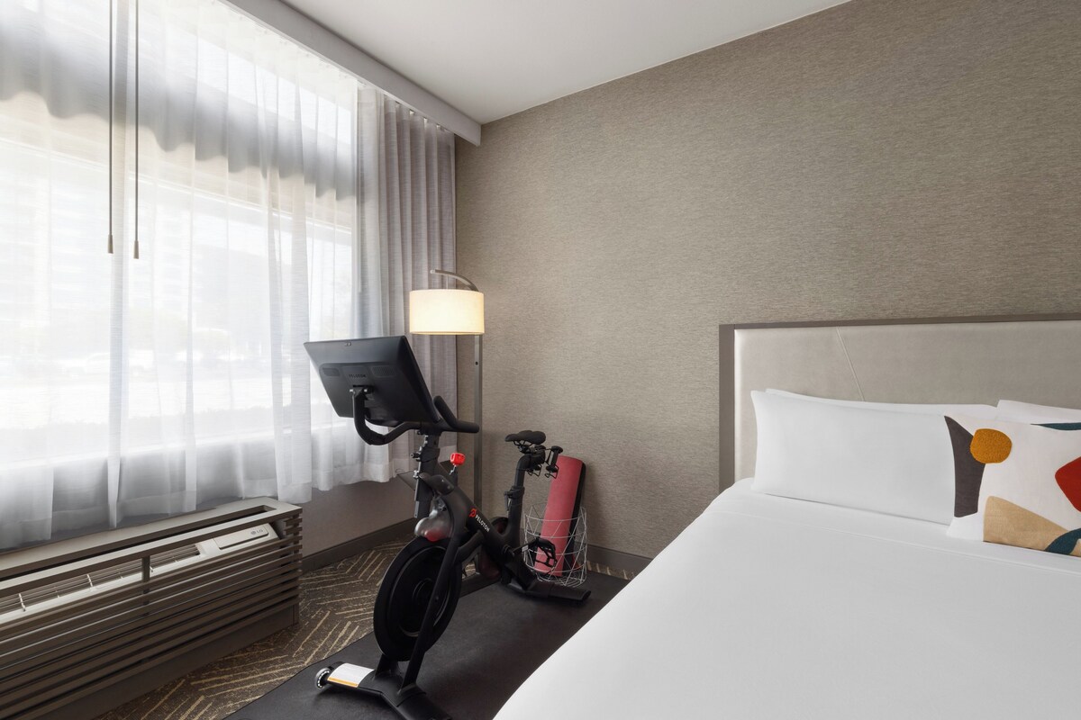 A hotel room with a bed, a desk with a laptop, a standing desk, and a stationary bike.