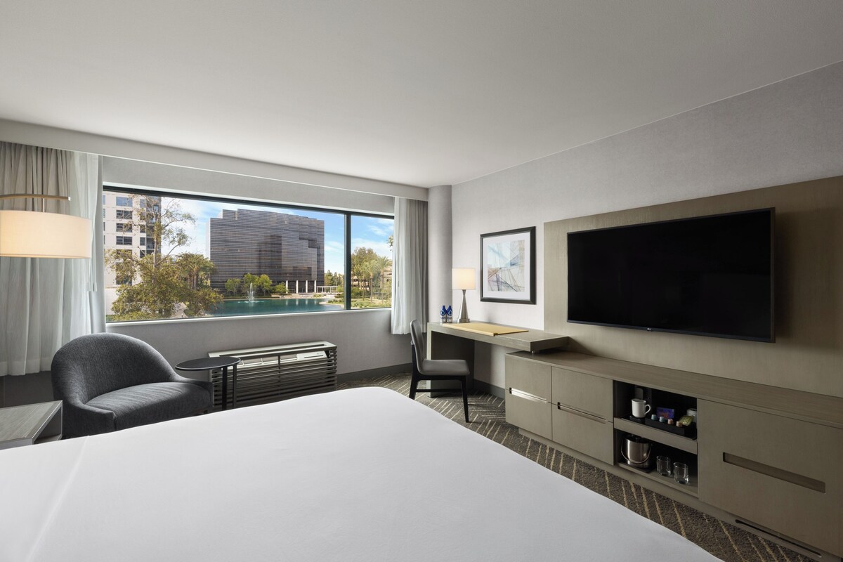 A hotel room with a bed, a desk, a chair, a TV, and a view of a building and trees.