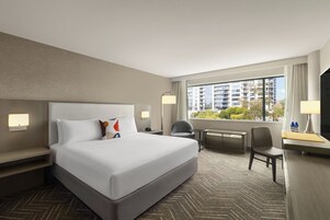 A hotel room with a large bed, a desk, a chair, and a view of a cityscape through the window.