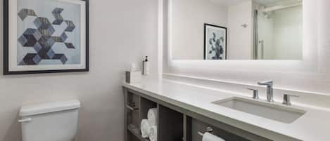 Un baño moderno con inodoro, lavamanos, espejo y ducha. Hay toallas y artículos de aseo en la encimera.